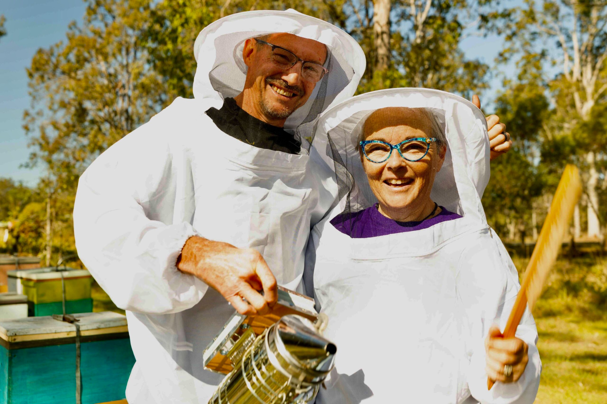 Two beekeepers smiling together beside their hives, holding a smoker and hive tool